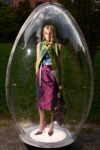 Woman wearing a lime green vest, blue top, and magenta skirt stands inside a clear glass capsule outdoors, holding a brown bag and wearing purple heels.