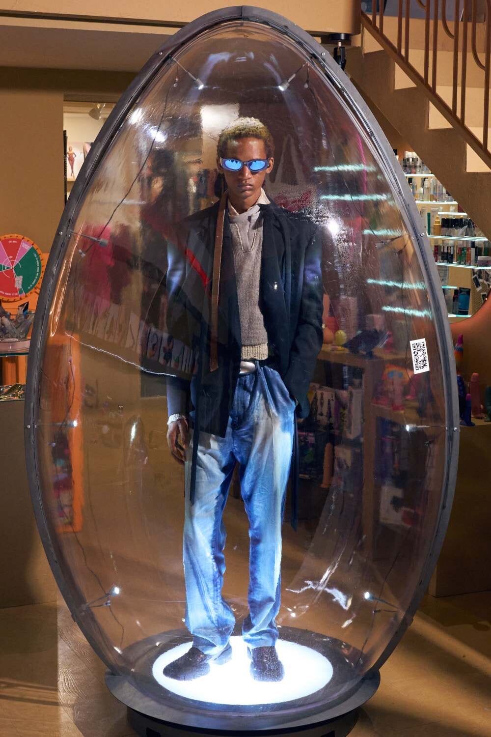 Man wearing sunglasses, blazer, and light sweater stands inside a clear egg-shaped display pod in a store window.