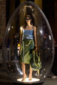 Model standing inside a large clear bubble on a lit pedestal, wearing sunglasses, a blue sleeveless top and a green skirt.