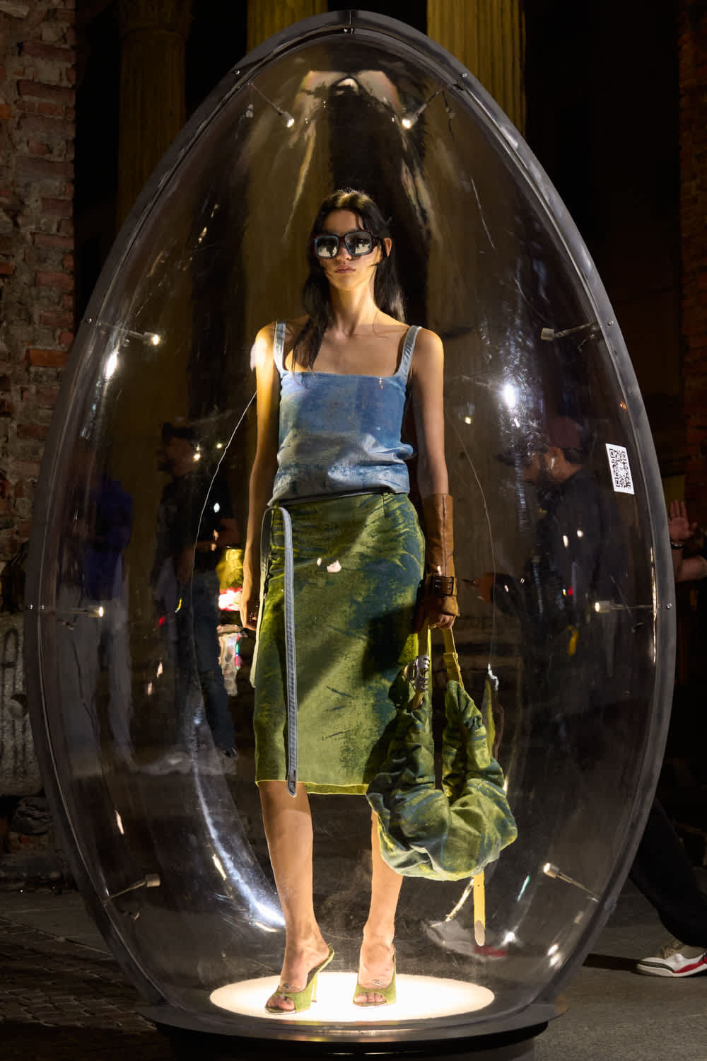 Model standing inside a large clear bubble on a lit pedestal, wearing sunglasses, a blue sleeveless top and a green skirt.