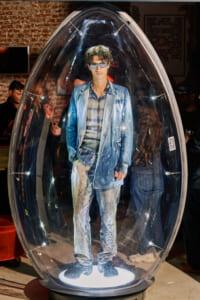 Man in a blue denim suit and sunglasses stands inside a clear egg-shaped capsule on a lit pedestal at an exhibition.