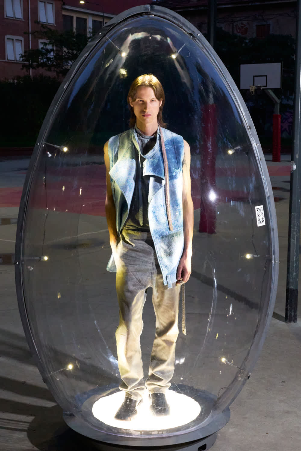 Person standing inside a clear oval pod illuminated from below, wearing a blue sleeveless jacket and beige pants.