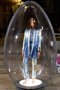 Model in a light blue denim suit stands inside a clear egg-shaped display pod at night, holding a small beige purse.