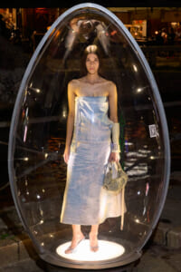 Model in a pale blue strapless dress standing inside a transparent egg-shaped display capsule at night, holding a woven bag.
