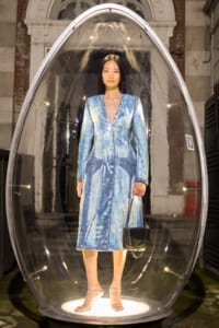 Model in a blue tie-dye dress standing inside a clear oval capsule, holding a black handbag on a runway-style setup.