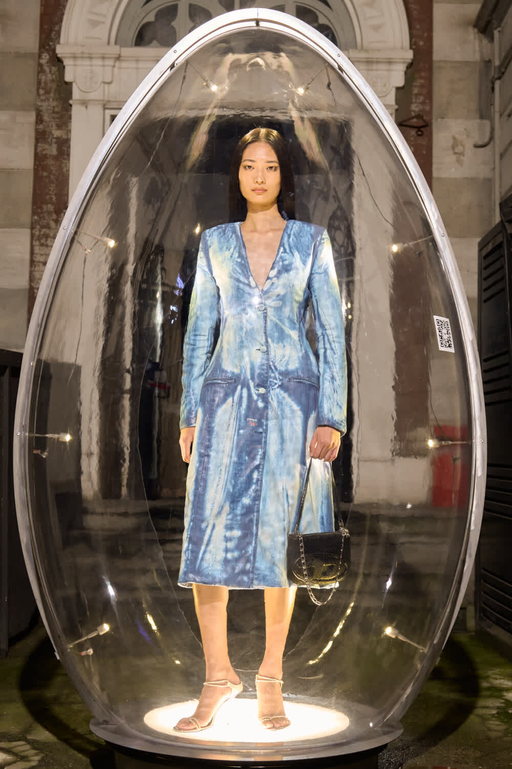 Model in a blue tie-dye dress standing inside a clear oval capsule, holding a black handbag on a runway-style setup.
