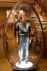 Man in a colorful shirt and jeans stands inside a clear, egg-shaped glass pod with a bright base light in a store window display.