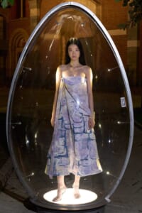 A model stands inside a clear teardrop-shaped glass capsule at night, wearing a lavender strapless dress and sandals.