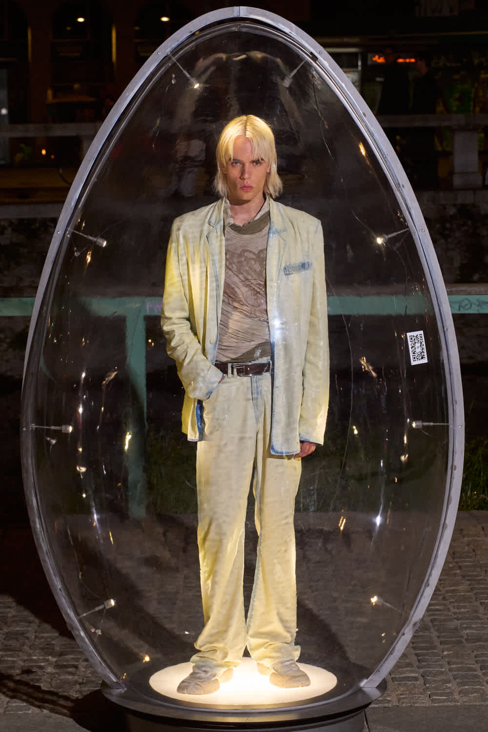Person with blond hair stands in a pale yellow velvet suit inside a clear, egg-shaped glass display illuminated from below at night.