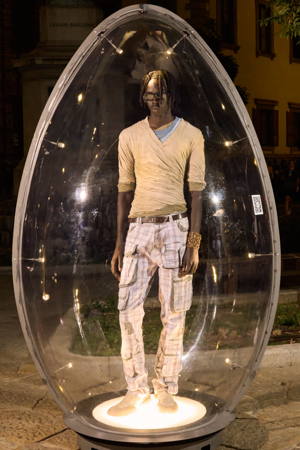 Man stands inside a clear oval capsule on a glowing pedestal, an urban night installation art piece behind him.