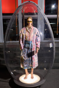 Person standing inside a clear egg-shaped capsule on a pedestal, wearing a colorful oversized coat, denim skirt, high heels, and yellow sunglasses.