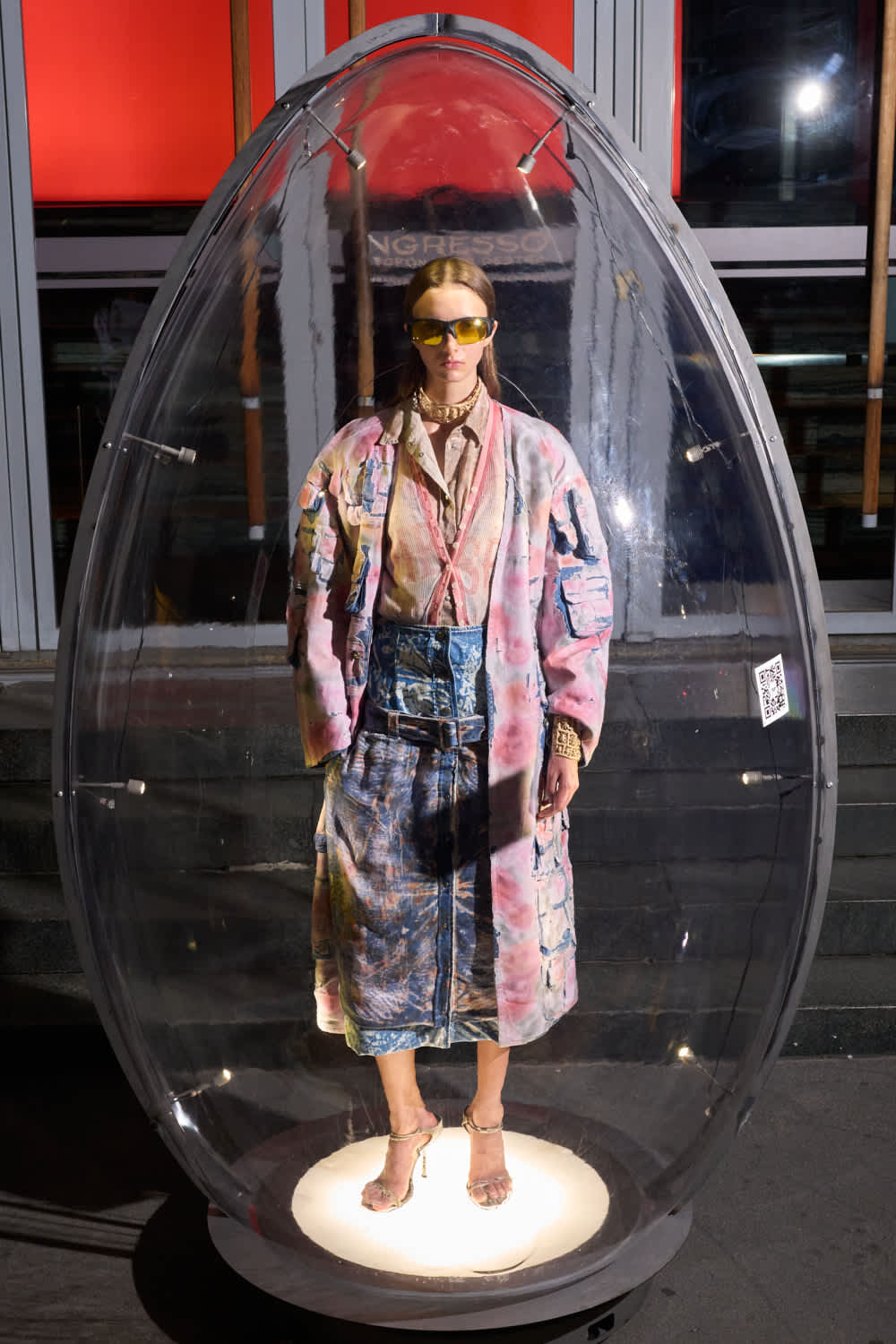 Person standing inside a clear egg-shaped capsule on a pedestal, wearing a colorful oversized coat, denim skirt, high heels, and yellow sunglasses.