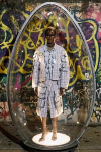 Person wearing a light plaid jacket and shorts stands inside a clear pod against a colorful graffiti wall behind him.