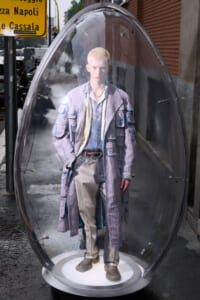 Life-size mannequin in a distressed lavender patchwork coat and beige pants inside a clear glass capsule on a city sidewalk.
