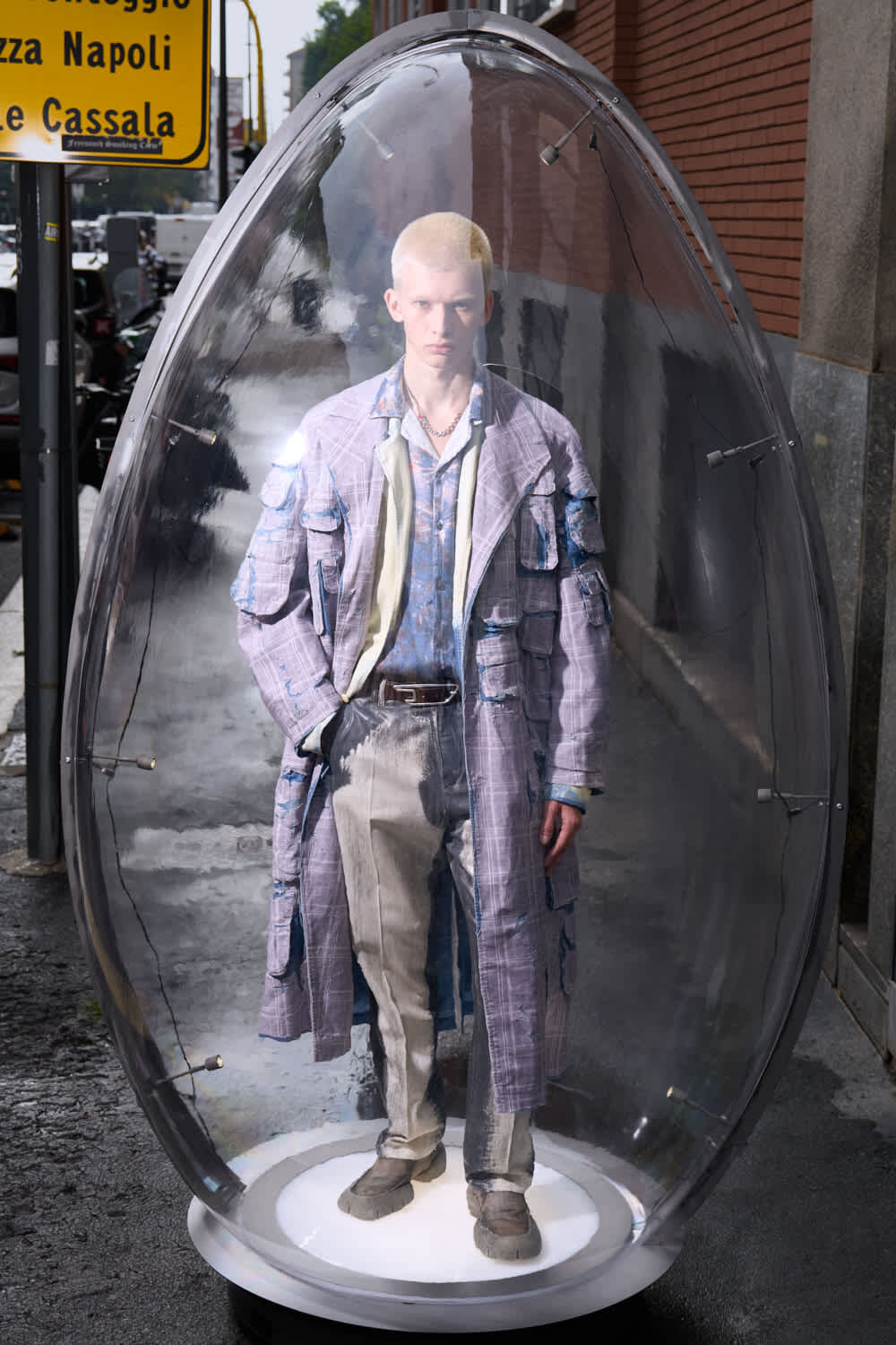 Life-size mannequin in a distressed lavender patchwork coat and beige pants inside a clear glass capsule on a city sidewalk.