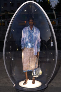 Person stands inside a glass pod on a city street at dusk, wearing a blue denim jacket and a beige skirt, holding metallic bags.