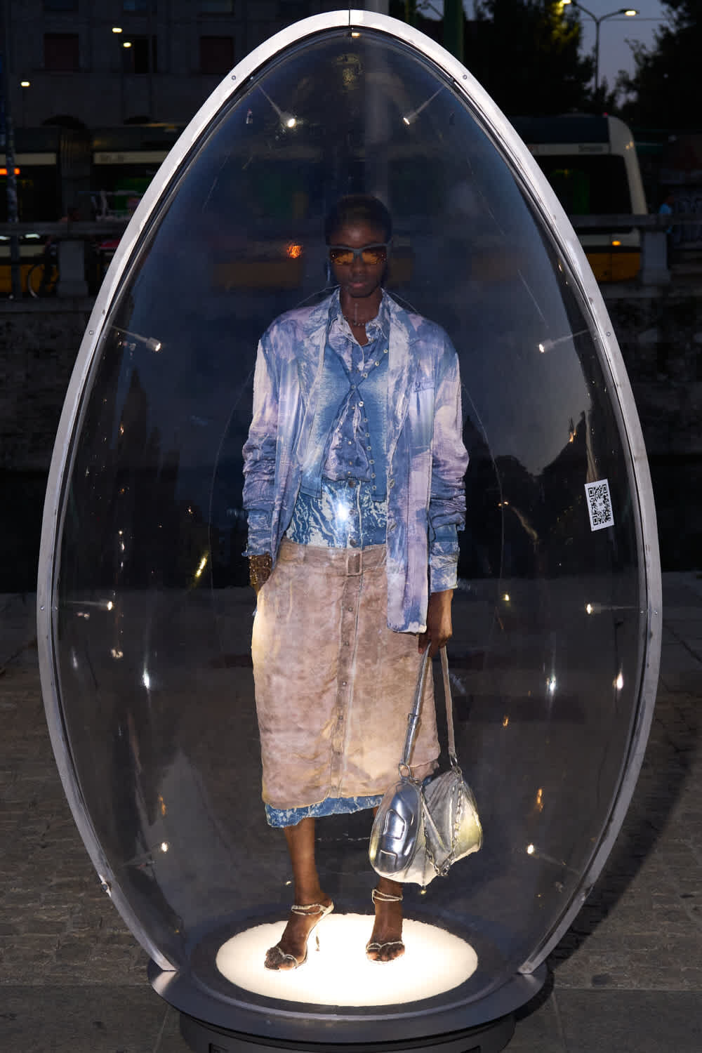 Person stands inside a glass pod on a city street at dusk, wearing a blue denim jacket and a beige skirt, holding metallic bags.