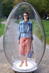 Model standing inside a clear capsule outdoors, wearing a denim jacket, pink midi skirt, sandals, sunglasses, and a blue handbag.