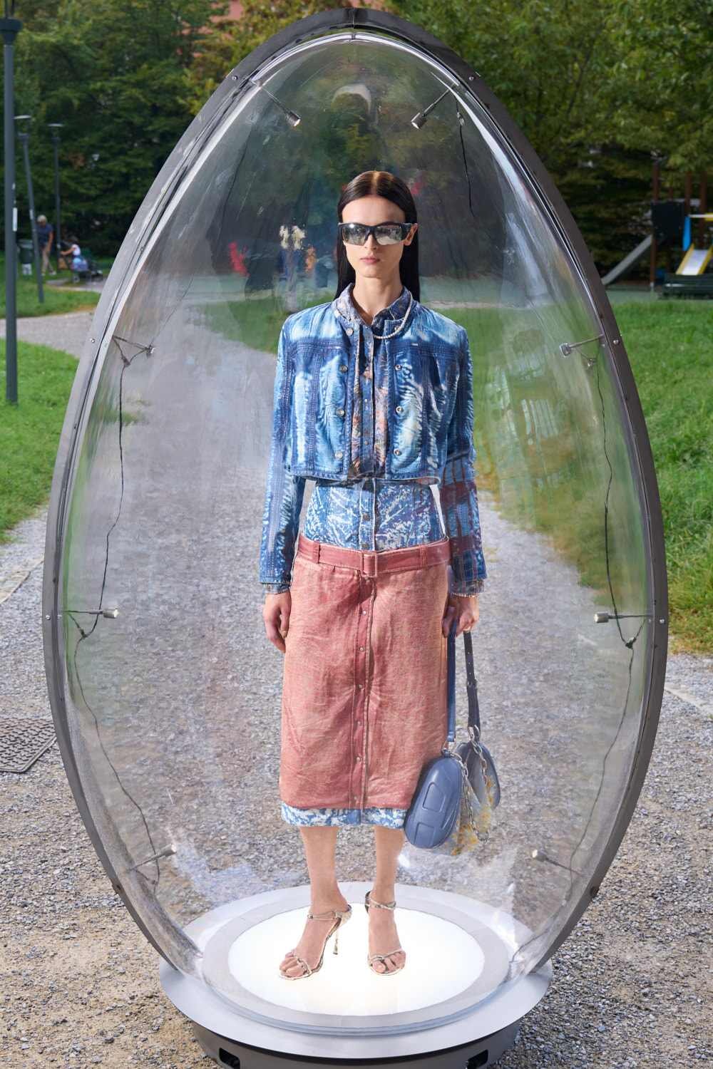 Model standing inside a clear capsule outdoors, wearing a denim jacket, pink midi skirt, sandals, sunglasses, and a blue handbag.