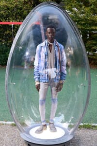 Young man stands inside a large transparent egg-shaped capsule on a playground, wearing a blue denim jacket over a grey sweater and light jeans.