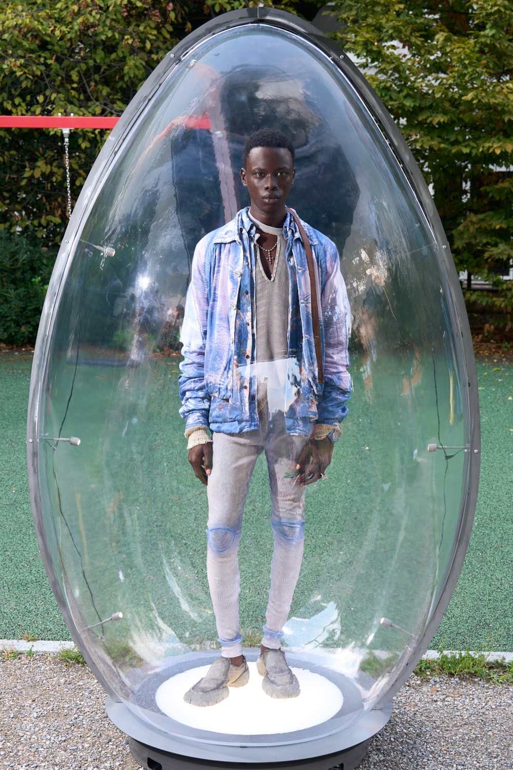 Young man stands inside a large transparent egg-shaped capsule on a playground, wearing a blue denim jacket over a grey sweater and light jeans.
