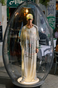 Man wearing a long beige robe stands inside a clear bubble-like pod on a street, looking at the camera.