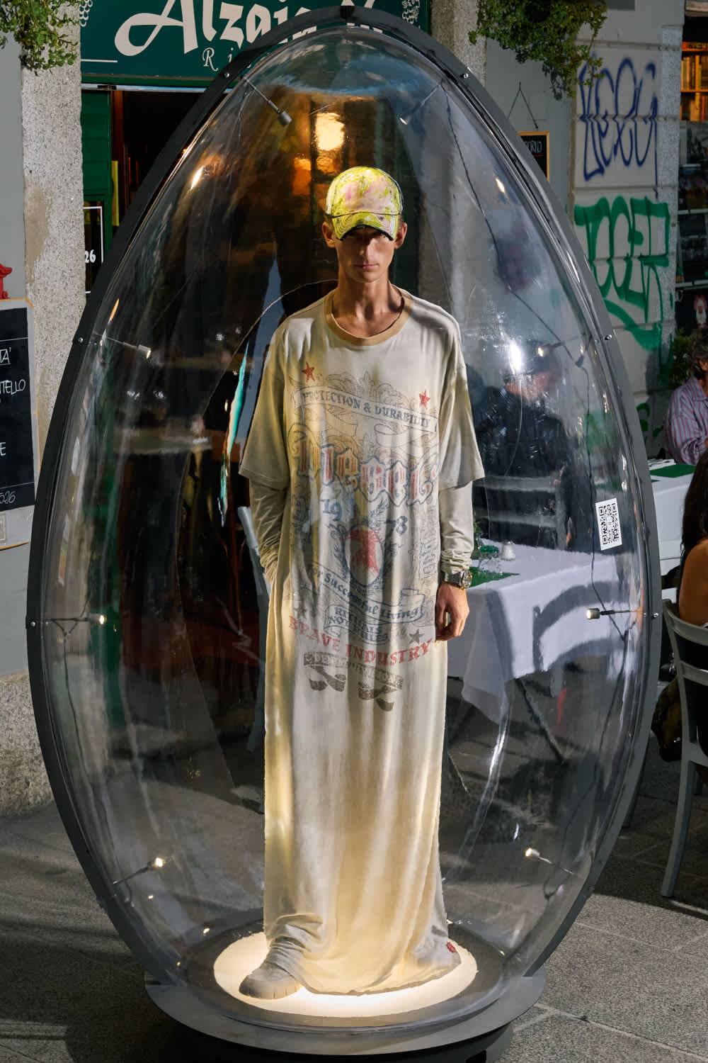 Man wearing a long beige robe stands inside a clear bubble-like pod on a street, looking at the camera.