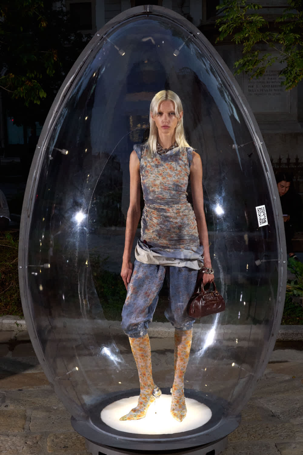 Model with blonde hair stands inside a clear capsule sculpture on a lit pedestal, wearing a blue-gray patterned outfit and holding a brown handbag.