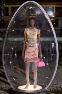 Model inside a transparent oval capsule on a pedestal at night, wearing a beige textured top and pink skirt with a pink handbag.