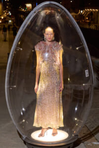 Model standing inside a transparent teardrop capsule on a city sidewalk at night, wearing a long pastel floral dress and high heels.