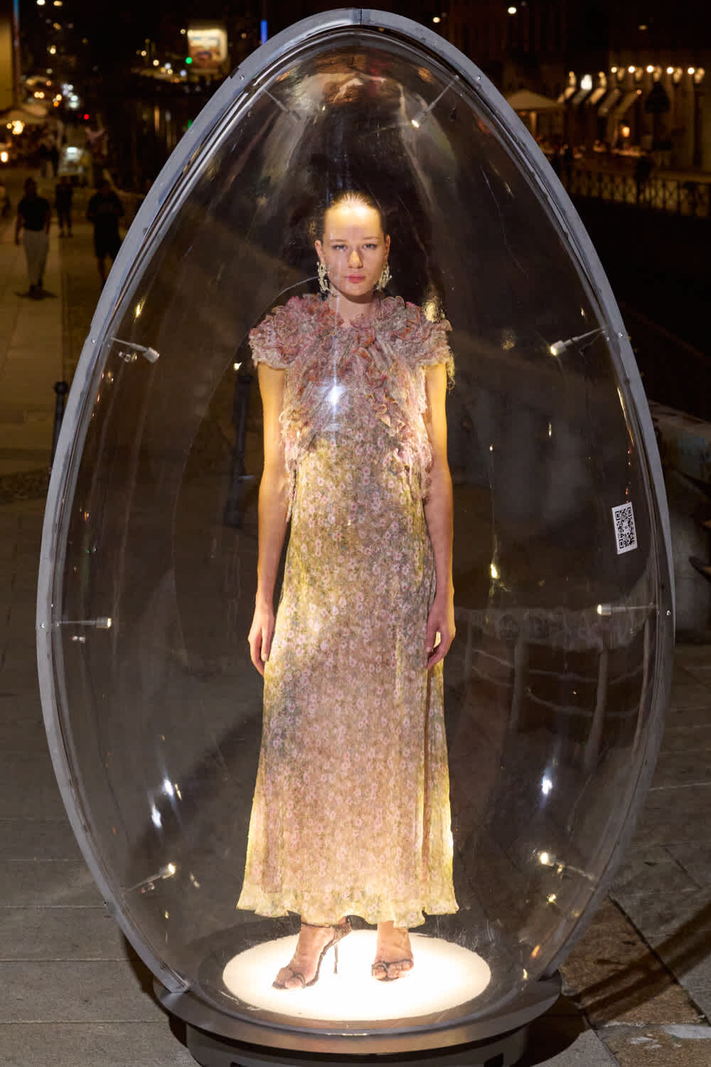 Model standing inside a transparent teardrop capsule on a city sidewalk at night, wearing a long pastel floral dress and high heels.