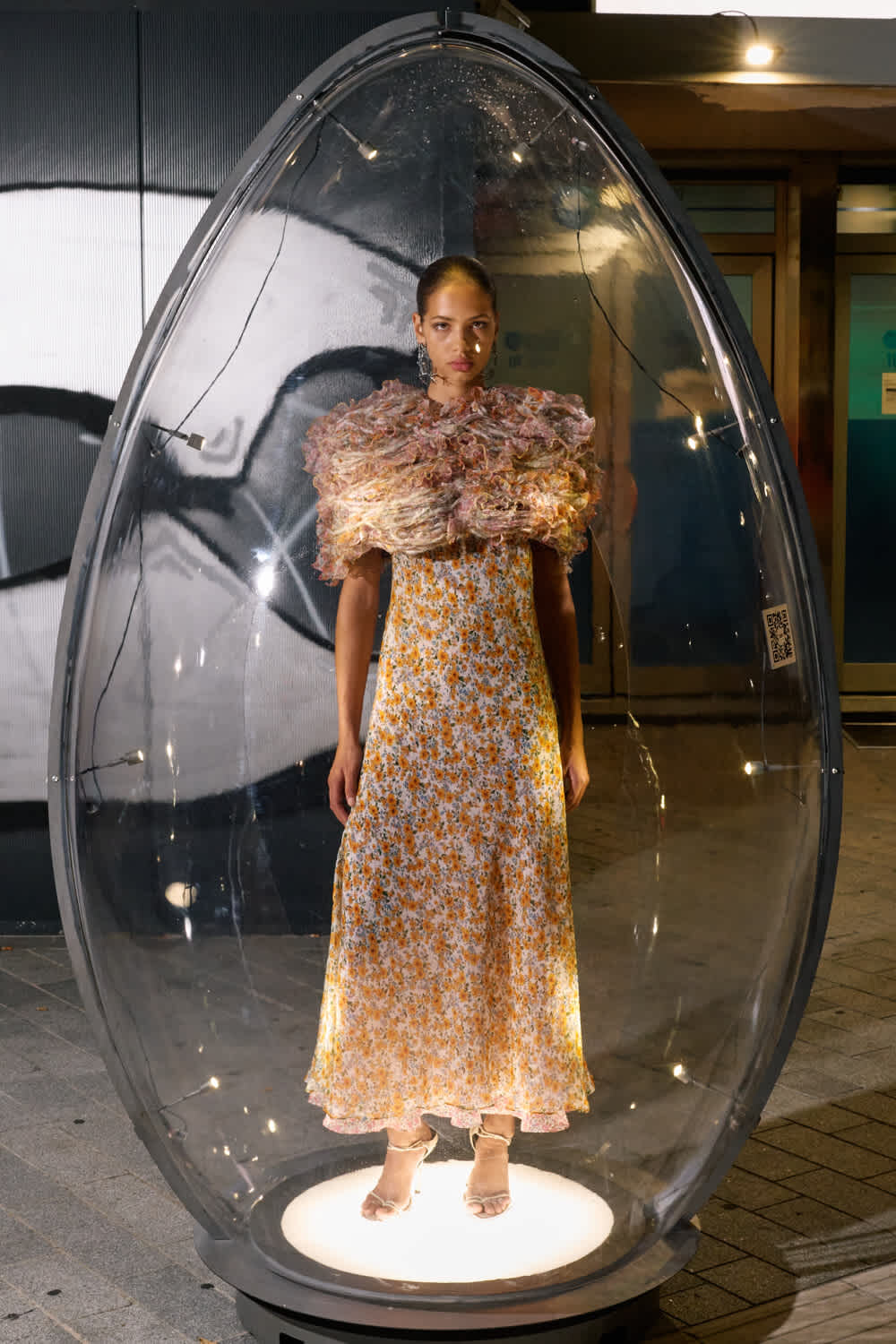 Model wearing a yellow floral maxi dress with a pink, ruffled collar inside a transparent, egg-shaped display capsule at a fashion exhibit.