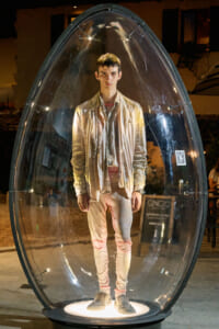 Young man stands inside a transparent capsule on a circular lit base at night, wearing pale jacket and pants.