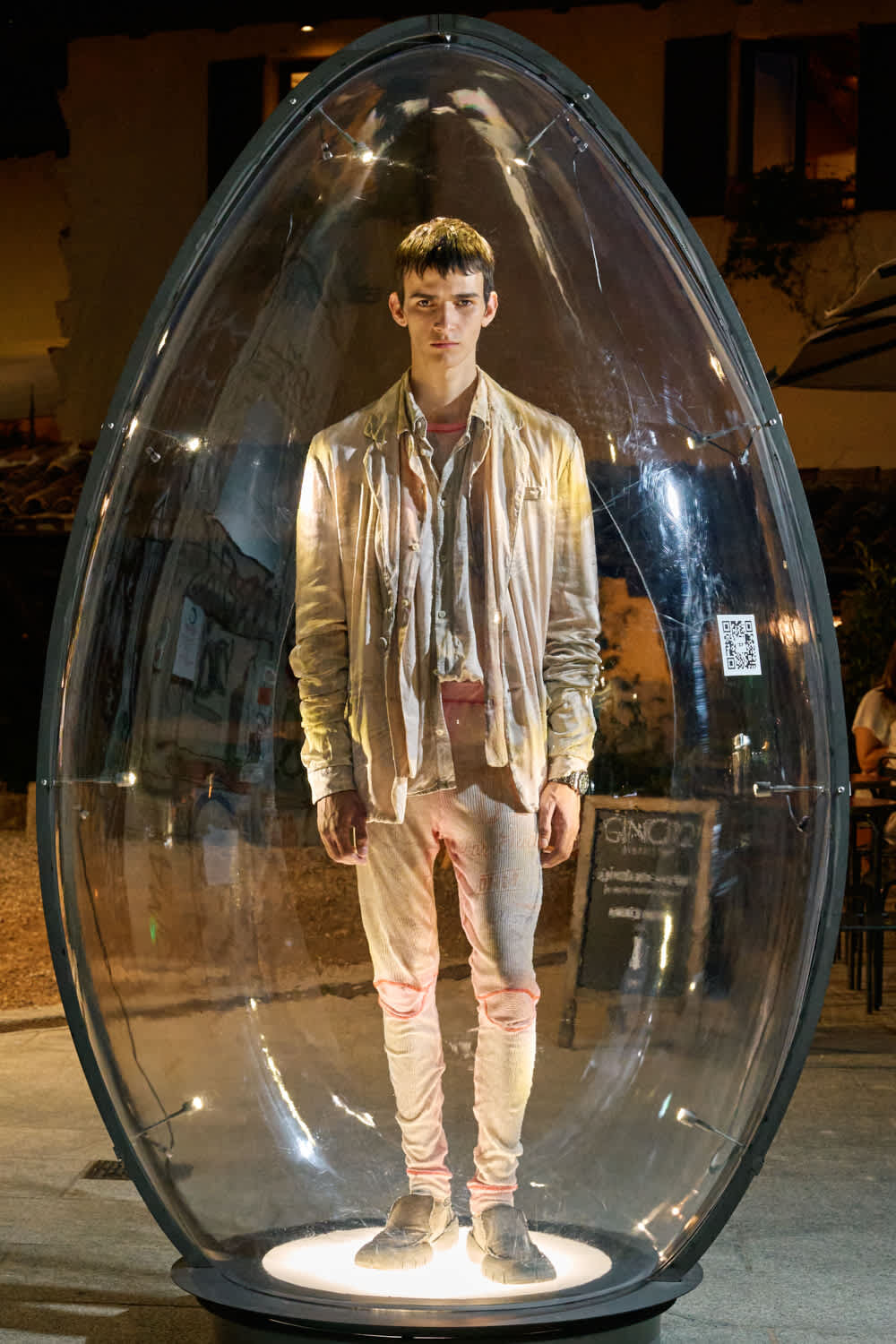 Young man stands inside a transparent capsule on a circular lit base at night, wearing pale jacket and pants.