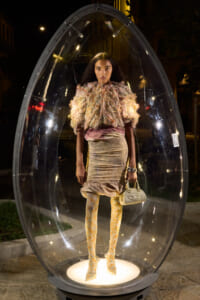 Model poses inside a clear egg-shaped display at night, wearing a voluminous textured ruffled top, a pale pink skirt, yellow patterned tights, and carrying a small white handbag.