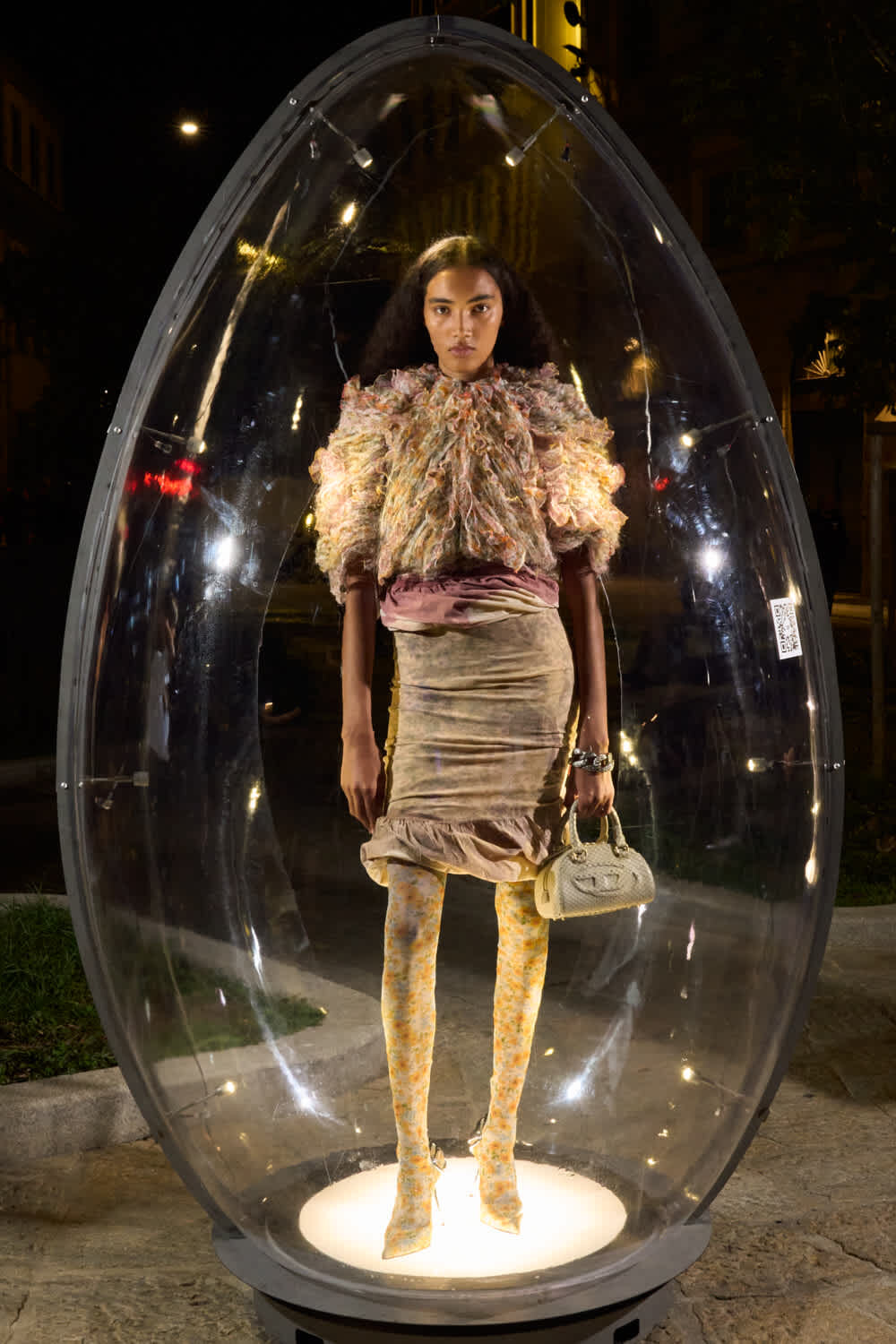 Model poses inside a clear egg-shaped display at night, wearing a voluminous textured ruffled top, a pale pink skirt, yellow patterned tights, and carrying a small white handbag.