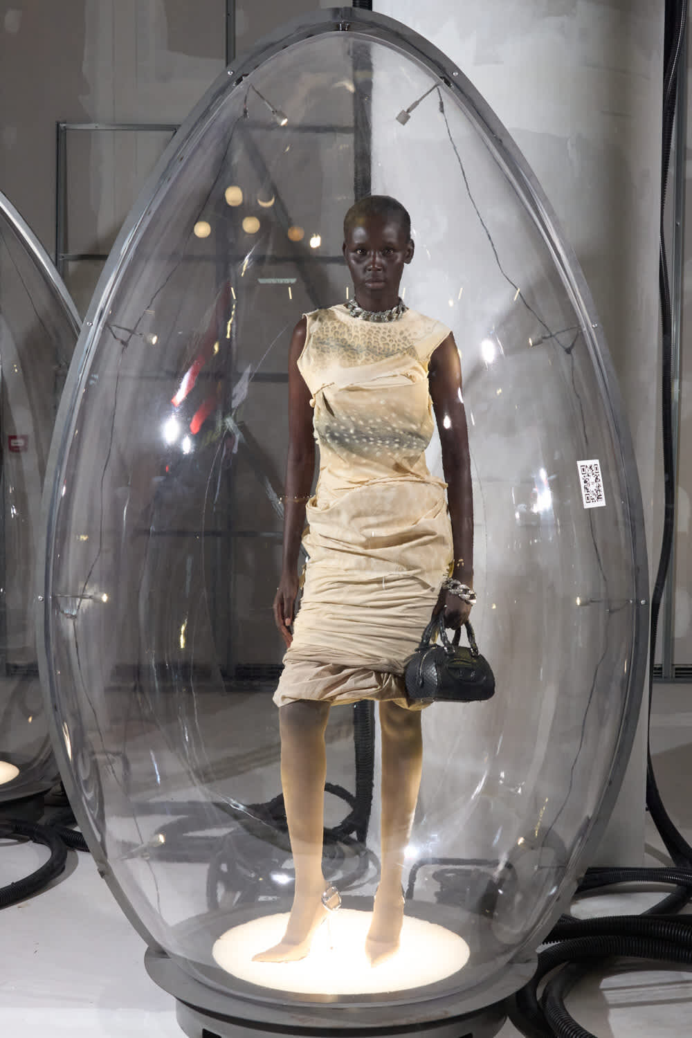 Model in a beige ruched dress stands inside a tall clear glass pod at a fashion exhibit, holding a black handbag.