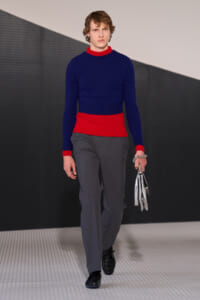 Male model walking a runway in a navy blue knit sweater with red neck and hem trim, gray trousers, black shoes, and holding a silver metallic clutch.