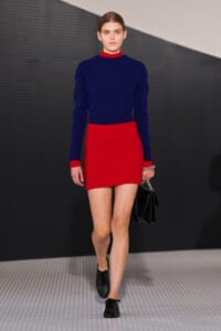 Model walking a runway in a cobalt blue knit sweater and red mini skirt, carrying a black handbag with a chain strap.