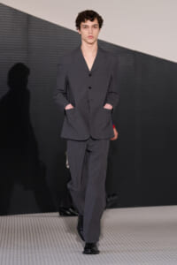 Male model wearing a dark gray suit with hands in pockets walking on a runway.