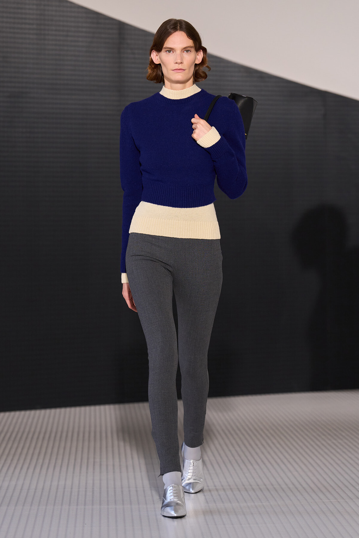 Female model walking a runway in a royal blue knit sweater with cream cuffs and hem, gray fitted pants, silver shoes, and a black shoulder bag.