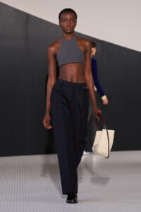Model walks the runway in a gray halter crop top and navy tailored pants, carrying a cream tote with brown handles.