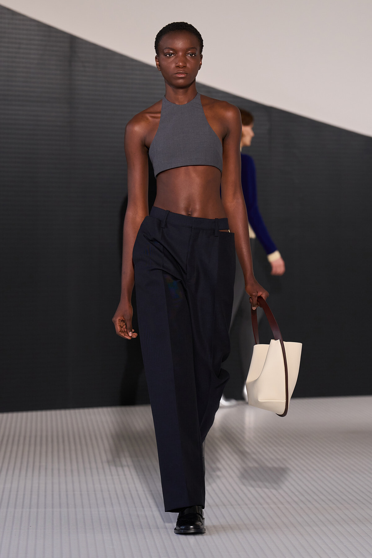 Model walks the runway in a gray halter crop top and navy tailored pants, carrying a cream tote with brown handles.