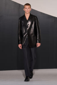Male model wearing a shiny black leather blazer over dark trousers, walking on a fashion runway.