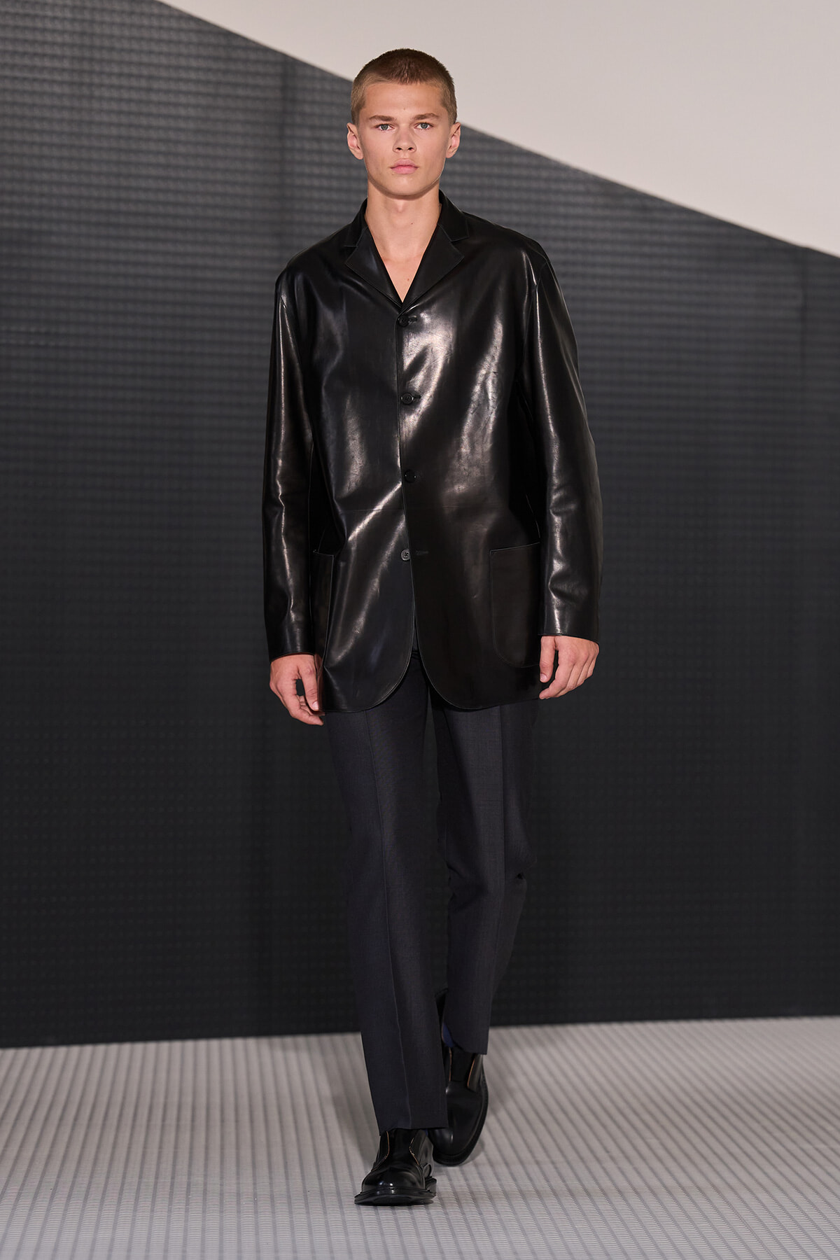 Male model wearing a shiny black leather blazer over dark trousers, walking on a fashion runway.