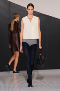 Runway model in a minimalist white sleeveless top, gray shorts, navy tights, black shoes, carrying a black handbag with burgundy handles.