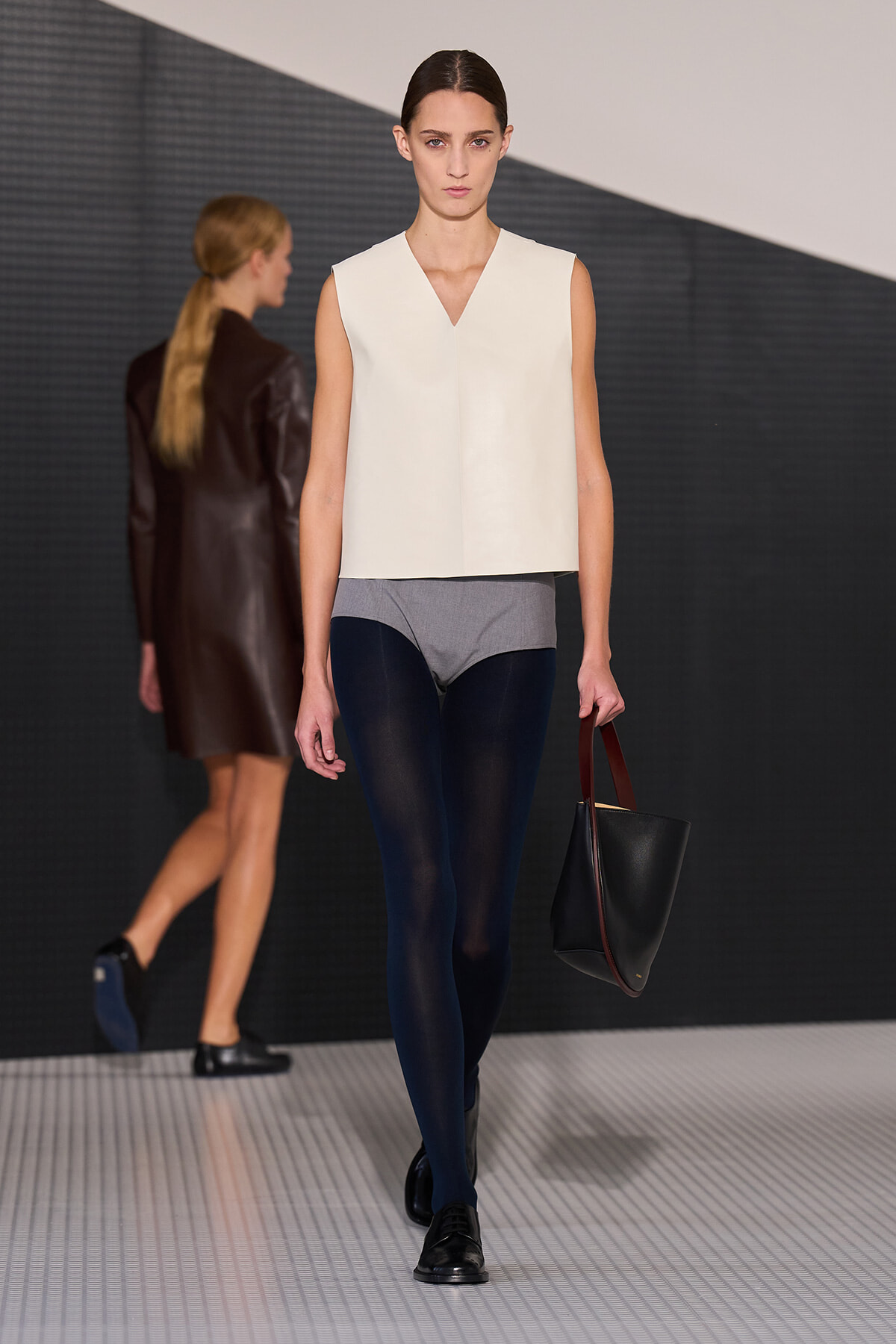 Runway model in a minimalist white sleeveless top, gray shorts, navy tights, black shoes, carrying a black handbag with burgundy handles.