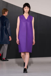 Model walking the runway in a sleeveless, violet V-neck shift dress with black loafers.