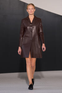 Female model walks a runway in a knee-length dark brown leather coat with a single-breasted button closure.