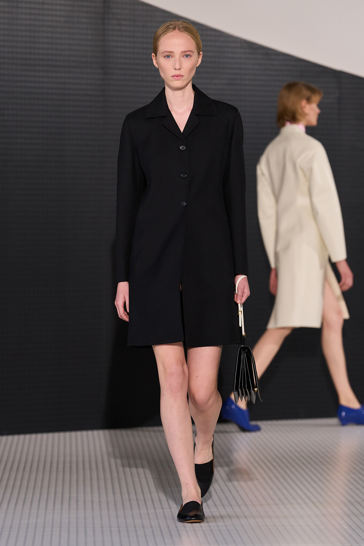 Model walking a runway in a black knee-length coat with a button front, carrying a small black clutch; another model in a cream coat is in the background.